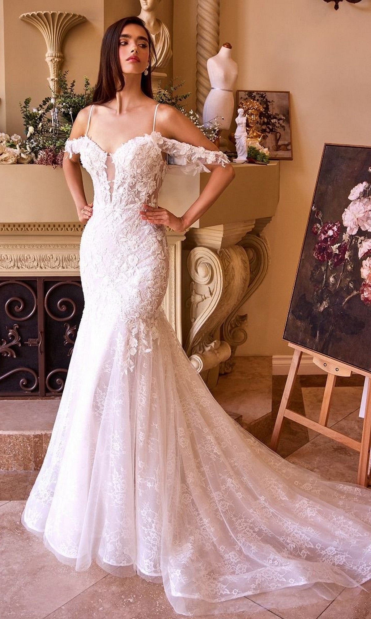 A woman wearing the Andrea and Leo WL051 long wedding dress, an off-the-shoulder fitted mermaid gown with white lace appliqué and a long train, stands indoors near a fireplace and floral painting.
